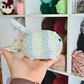 A hand holding a crocheted toy cat in front of a shelf filled with colorful yarn balls.