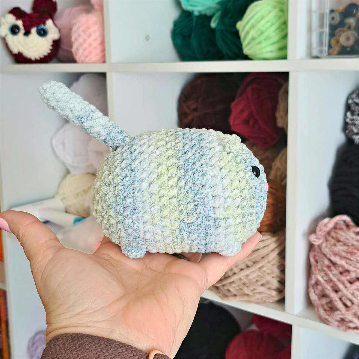 A hand holding a crocheted toy cat in front of a shelf filled with colorful yarn balls.