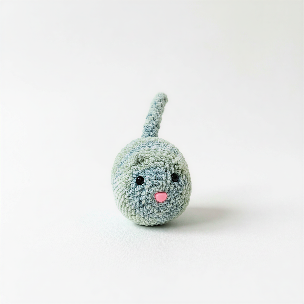 A small, crocheted toy cat with a pink nose and eyes sits on a plain white background.