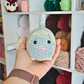 A hand holding a small, crocheted toy resembling a cat with a pink nose, in front of a shelf filled with colorful yarn balls.