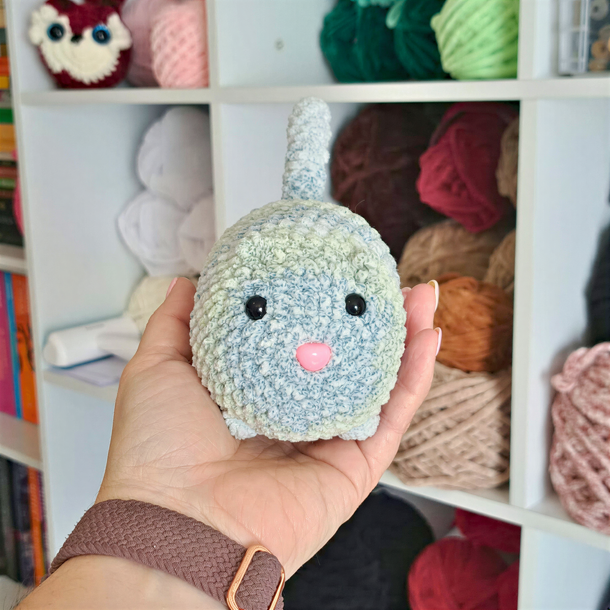 A hand holding a small, crocheted toy resembling a cat with a pink nose, in front of a shelf filled with colorful yarn balls.