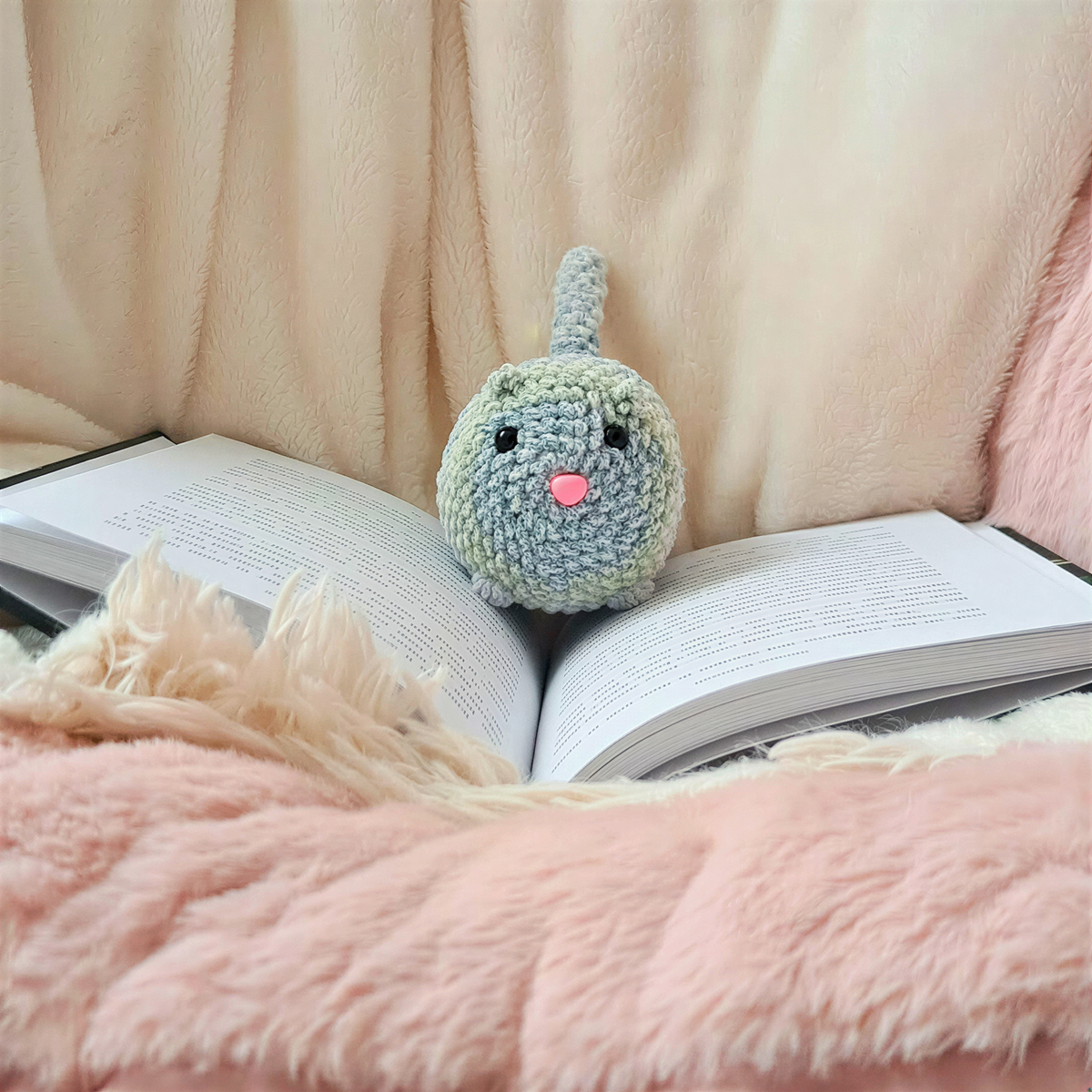 A crocheted cat toy with a pink nose sits on an open book, surrounded by a cozy pink blanket.